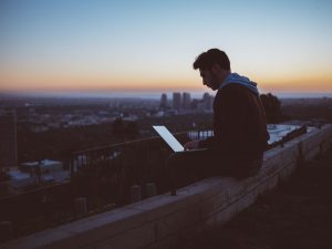 man sat on a ledge on a laptop infront of a sunset